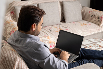 Shoulder view young indian male holding laptop computer mock up blank white screen on sofa. Man using laptop surfing internet, read news, distance online office work concept advertising work from home