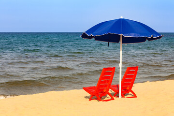 Red beach loungers and blue umbrella set on sandy shore with sea view