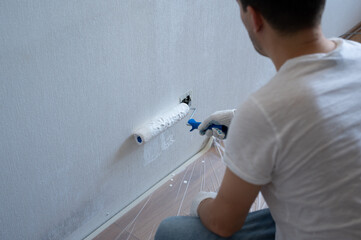 Painter working using roller and brush painting white wall near power socket during home renovation