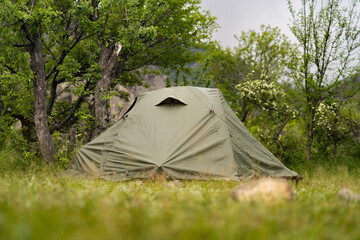 Green tent at a secluded campsite in green forest in mountains with cloudy sky in the background, place for tent outdoor, tourism and summer holidays concept