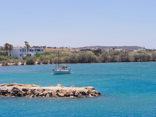 Scenic coastal view with turquoise water, rocky shore, white buildings, and a sailboat in the distance. Punta port, Paros island, Greece