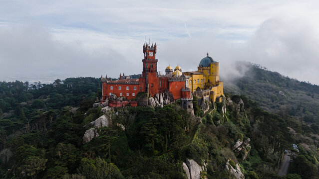 Aerial view of the vibrant Pena Palace perched atop a hill amidst lush greenery, a striking contrast against the misty sky, Sintra, Lisboa, Portugal.