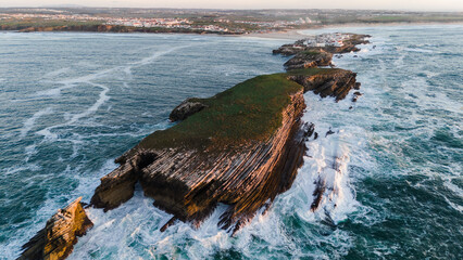 Aerial view of rugged cliffs meet the turbulent ocean at Baleal Island, the waters churning with white foam against the dark rock, Baleal, Leiria District, Portugal.