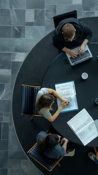 An aerial perspective showcases a group of colleagues working together at a round table. Laptops and documents are scattered around, emphasizing cooperation in a modern office environment.