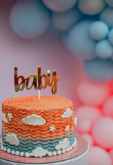 Bright baby shower cake with “baby” topper, orange-blue color scheme, and pastel balloon decor in the background. Playful and joyful celebration moment.