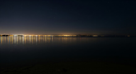 City lights reflecting on the calm water at night under a starry sky