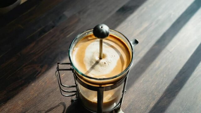 A top-down shot of hot coffee with a rich crema brewing in a glass French press on a wooden table, with strong sunlight creating a high-contrast scene
