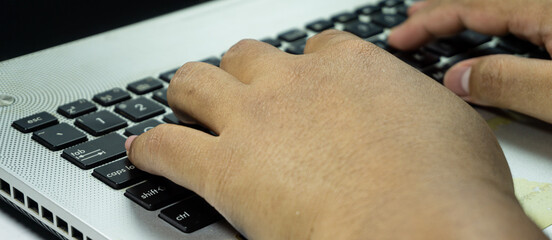Indonesian man’s hand typing on laptop keyboard with black keys and silver case, close-up shot...