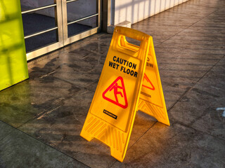 A bright yellow "Caution Wet Floor" sign stands on a shiny tiled surface, illuminated by warm sunlight. This safety sign is a common sight in public spaces, reminding people to be careful in slippery
