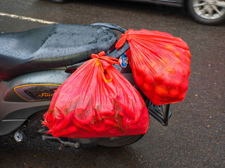 Two large red plastic bags filled with chili peppers and tomatoes are tied to the back of a motorcycle on a wet street. A real-life glimpse into traditional market transportation in urban settings.