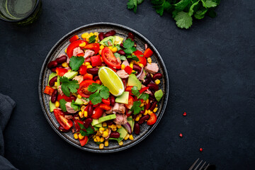 Tex-mex salad with canned tuna, avocado, corn, red beans, cherry tomatoes, cilantro and lime dressing, black table background, top view