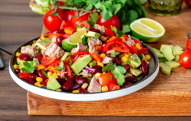 Preparing yummy salad with tuna, corn, avocado, beans, tomatoes and cilantro in plate on kitchen countertop and ingredients for preparation on wooden cutting board