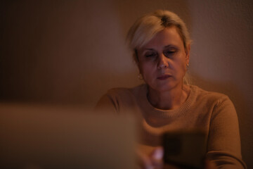 Mature woman in a dimly lit room looking at her smartphone, wearing a beige sweater. The atmosphere...