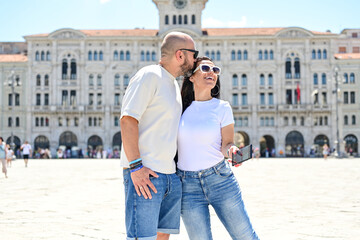 Obraz premium Trieste ,Italy, Piazza Unita` .Happy smiling couple of tourists making photos near Piazza Unita` at Trieste .Concept of Italian gastronomy and travel. Italian couple having fun together