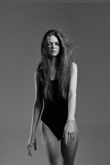 black and white woman with long hair wearing black bodysuit standing against gray background in...