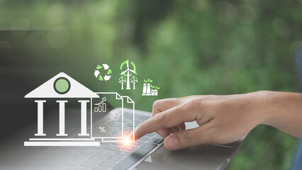 Close-up of a hand typing on a laptop with green finance icons, including bank, wind turbines, recycling, and eco industry, symbolizing sustainable investment and digital environmental economy.