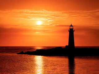 Silhouette of a lighthouse against a deep orange sunset sky with relecttions in the sea. No people. Travel concept.