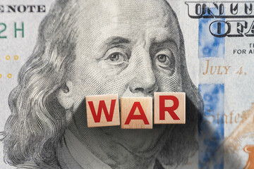 Wooden blocks spelling “WAR” placed on a U.S. dollar bill with Benjamin Franklin’s portrait,...