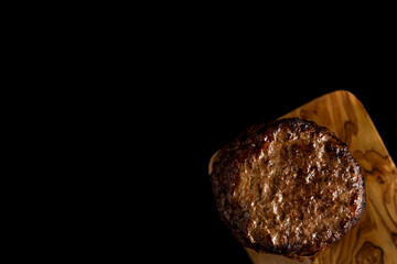 A single, perfectly cooked and juicy burger patty rests on a small, rustic wooden cutting board against a dark background