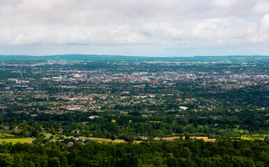 A vast cityscape stretching into the distance, seen from an elevated perspective, with lush green trees and fields dominating the foreground under a bright cloudy sky