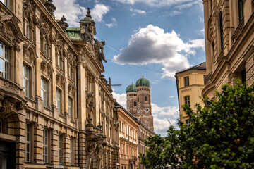 Iconic Cathedral Of Our Dear Lady In Munich: Majestic Twin Domes Rising Above Historic Buildings Under A Bright Blue Sky With Scenic Clouds Near Marienplatz