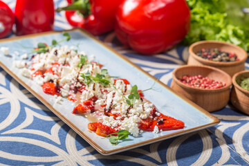 The photo shows a rectangular blue plate with a colorful dish featuring roasted red bell pepper slices topped with crumbled white cheese, likely feta, and garnished with microgreens. 