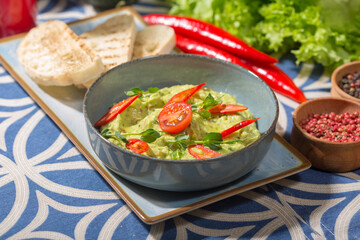 The photo shows a blue bowl filled with a creamy green dip, likely guacamole, garnished with red chili slices, a cherry tomato, and fresh microgreens. 