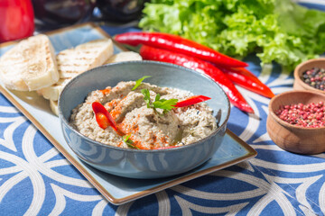The photo shows a blue bowl filled with a creamy dip, likely baba ganoush or hummus, garnished with red chili slices, parsley, and a sprinkle of spices and sesame seeds.