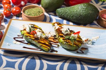 two halves of a grilled avocado, each topped with crumbled blue cheese, almond slices, a sprig of rosemary, and a small red chili slice