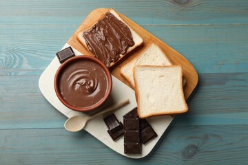 Tasty chocolate butter, pieces, spoon and slices of bread on light blue wooden table, top view