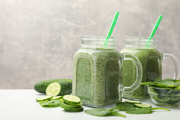 Tasty smoothie in mason jars, spinach leaves and cucumber on white table against grey background,...