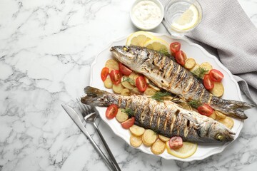 Tasty roasted seabass fish served with potatoes, tomatoes, sauce and lemon on white marble table, flat lay and space for text. Homemade seafood dish