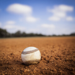 Worn baseball resting on the dirt of an empty field under a blue sky with fluffy white clouds.