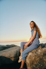 Lifestyle woman in modern boho-western denim street style sits on rocks by the ocean at sunset, captured with warm film color tones for a relaxed vibe.