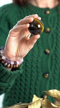 Extreme close-up video of a hand in an emerald green chunky knit sweater holding a rotating tiger's eye crystal sphere, adorned with natural stone bracelets. Bright white minimalist background with