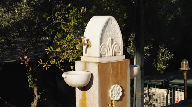Functioning Greek-style marble fountain near the Odeon of Herodes Atticus in Athens. Features ornate spigot, shell carving, floral motifs, and basin. Surrounded by greenery. No people 
