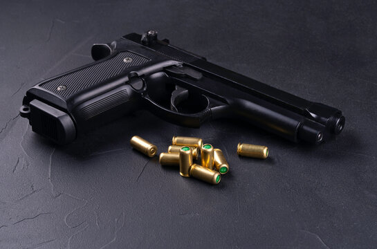 Black semi-automatic pistol with a textured grip lies on a dark surface next to several gold-colored bullets. The handgun is positioned at an angle, clearly displaying its barrel and trigger. This ima