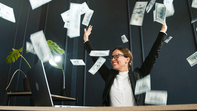 A successful middle-aged businesswoman enjoys a stack of money from a successful deal and closed contract in her office. Perfect for themes of business success, financial achievement.