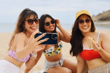Group of young Asian women taking a selfie at the beach while enjoying pizza. Happy friends smiling and having fun during a summer vacation by the sea. Friendship, travel, and lifestyle concept.