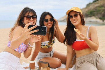 Group of young Asian women taking a selfie at the beach while enjoying pizza. Happy friends smiling...