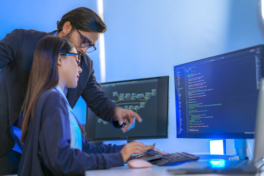 Asian female AI programmer and Indian male data analyst reviewing code on a monitor. Diverse tech professionals collaborating on an artificial intelligence project in a modern office.