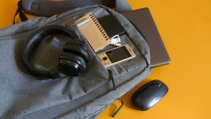 Modern study or travel setup featuring a roomy backpack with electronic gadgets—laptop, phone, powerbank, mouse, USB cable, notebook and cushioned headphones on yellow. High resolution
