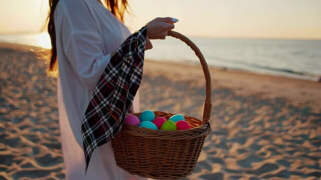 A woman carries a wicker basket with Easter Eggs, The setting sun and the wind blows the plaid on it. Sea sand and waves in the background