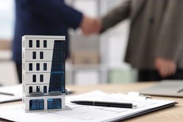 Real estate agent and client shaking hands at desk in office, focus on house model