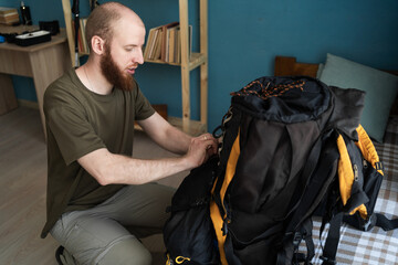Young bearded man traveler prepare for hiking vacation standing near bad with backpack and other...