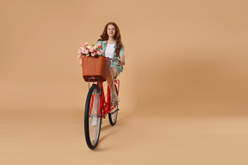 Beautiful teenage girl riding bicycle with basket of flowers against beige background. Space for...