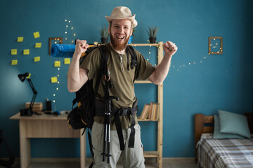Young bearded man traveler in hat prepare for hiking vacation at home smiling showing winner...