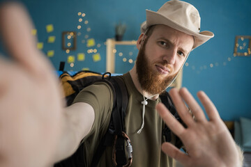 Nervous bearded traveler in hat taking selfie with negative facial expression and stop gesture no...