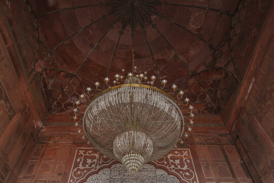 View of a grand chandelier hanging from a red ornate ceiling, casting a soft glow on the textured walls in New Delhi, Delhi Division, India.