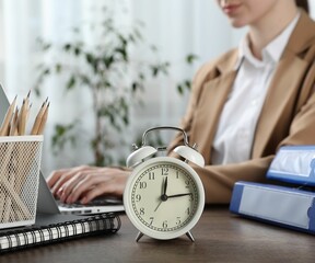 Deadline. Woman with laptop at wooden table in office, focus on alarm clock